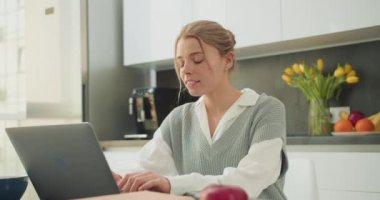 Genç bir kadın, parlak ve modern bir mutfakta dizüstü bilgisayarıyla çalışıyor. Sahne, taze çiçekler ve meyvelerin yer aldığı huzurlu bir ofis atmosferiyle üretkenlik ve dengeyi vurguluyor..