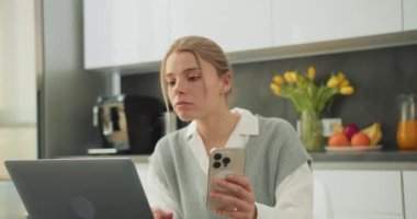 Modern mutfak ortamındaki bir kadın telefonuna odaklanmış endişeli ya da üzgün bir ifade sergiliyor. Bu resim dijital bir cihazla alınan bir endişe veya kötü haber anını iletir.