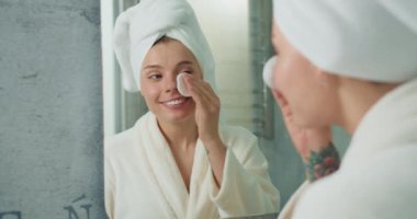 Cilt bakımı sırasında yüzüne pamuk pedi süren bir kadın. Havlu ve bornoza sarılmış halde banyo aynasının önündeki yüz bakımına odaklanıyor..