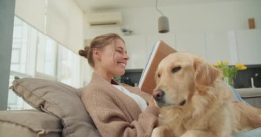 Genç bir kadın kitap okurken kanepede dinlenir ve Golden Retriever 'ı yanında dinlenir. Sıcak ve samimi sahne, arkadaşlığı, rahatlığı ve huzurlu bir ev atmosferini yakalar..