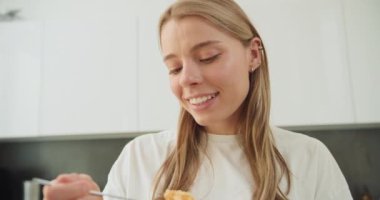 Modern bir mutfakta, kaşıktan mısır gevreği yiyen genç bir kadının yakın plan görüntüsü. Sağlıklı bir kahvaltıya daldığında, tipik bir zevk ve rahatlama ifadesi sergiler.
