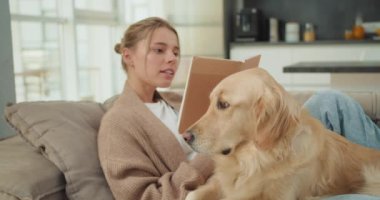 Genç bir kadın, modern bir oturma odasında kitap okurken Golden Retriever 'ıyla koltukta rahatça oturur. Sahne, rahatlama, arkadaşlık ve huzurlu bir atmosfer yakalar..