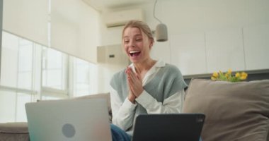 Bir kadın, iyi ışıklandırılmış bir oturma odasında otururken video görüşmesi sırasında şaşkınlık ve heyecanla sevinçle tepki verir. Parlak, çağdaş dekor canlı atmosfere ekleniyor.. 