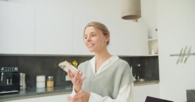 Modern bir mutfakta telefonuyla konuşurken gülümseyen bir kadın. Boşluğun temiz tasarımı, biraz dekor ile rahatlatıcı ve ilgi çekici bir sohbeti vurgular..