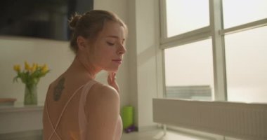 Bir kadın yoga pozunda sakince oturur, sakin yüz ifadesini ve sırt dövmesini vurgular. Sahne, modern bir odadaki büyük pencerelerden süzülen yumuşak güneş ışığıyla aydınlatılıyor..