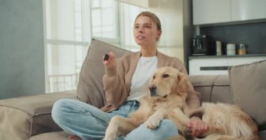 Genç bir kadın, yanında altın av köpeğiyle rahat bir kanepede oturuyor elinde uzaktan kumandayla, modern ve parlak bir oturma odasında eğlenmeye hazır..