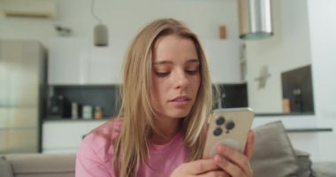 Pembe gömlek giyen genç bir kadın akıllı telefonunu kullanırken yoğun bir şekilde odaklanır. Minimalist dekorlu, parlak ve havadar bir atmosferi olan çağdaş bir oturma odasında oturuyor. İfadesi gösteriyor ki