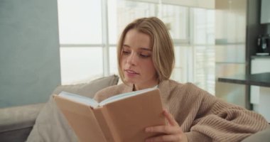 Genç bir kadın, büyük pencerelerden gelen doğal ışıkla dolu modern bir oturma odasındaki bir kanepede otururken kitap okuyarak huzurlu bir anın tadını çıkarıyor. Yüzü sakin ve odaklanmış görünüyor..