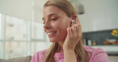 Genç bir kadın parlak, rahat bir oturma odasında akıllı telefonuyla iletişim kurarken kablosuz kulaklıklarla müzik dinlemekten mutlu olur. Sıradan giyiniyor ve rahatlama ortamının tadını çıkarıyor.