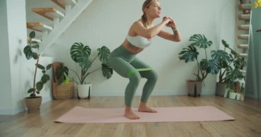 Kararlı bir kadın, yoga minderinde dengesini korumak için direniş bandıyla çömelme egzersizi yapıyor. Huzurlu, bitki dolu oda onun fitness rutinine barışçıl bir ortam ekler..