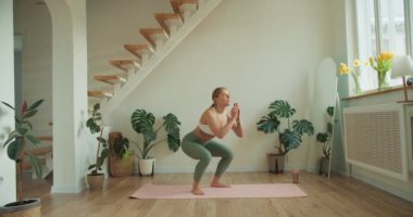 Bir fitness meraklısı sakin bir ev stüdyosunda çömelme gösterisi yapıyor. Yeşillik bitkiler ve yumuşak ışıklandırma ile minimalist düzenleme odağı artırır, güç ve denge duygusunu teşvik eder.