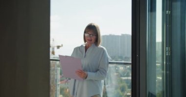 Olgun, profesyonel bir kadın elinde bir panoyla balkonda duruyor ve belgeleri inceliyor. Arka plandaki parlak şehir silueti üretken bir kurumsal ortamı yansıtıyor
