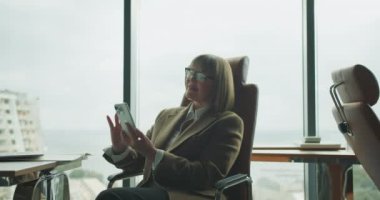 Spor ceket giymiş profesyonel bir kadın modern bir ofiste rahat bir şekilde akıllı telefonunu kullanır. Büyük pencereler, sakin bir çalışmanın altını çizerek şehir manzarası ve yeşillik manzarası sunuyor