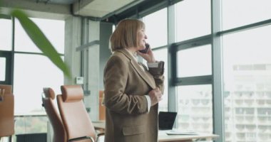 Kendine güvenen olgun bir iş kadını, modern bir ofiste büyük pencereleri ve parlak doğal ışığı olan bir telefon görüşmesi yaptı. Liderlik ve profesyonel iletişim temaları için ideal.