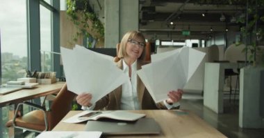 Modern ofis ortamında neşeli bir iş kadını başarıyı kutlamak için kağıtları havaya fırlatıyor. Arkaplandaki büyük pencereler şehir manzarasını gösterir.