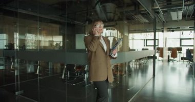 Modern bir ofiste kendine güvenen bir iş kadını elinde dosyalarla bir pano tutarken bir telefon görüşmesi yapıyor. Çevrede cam duvarlar ve çağdaş mobilyalar yer alıyor.