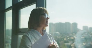 Büyük bir ofis penceresinin yanında elinde kalem ve not panosuyla duran gözlüklü olgun bir profesyonel kadın. Arka plandaki parlak şehir manzarası modern bir kurumsal ortamı yansıtır.