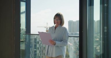 Profesyonel bir kadın cam bir balkonda duruyor ve iş belgelerini bir panoda gözden geçiriyor. hareketli bir şehir manzarasının arka planı modern şirket dünyasını ve liderliği vurguluyor.