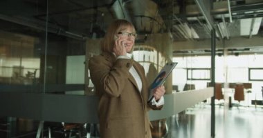 Modern bir ofisteki kendine güvenen bir iş kadını elinde veri tablosuyla bir telefon görüşmesi yapıyor. Cam bir bölmenin yanında duruyor, liderlik ve profesyonelliği somutlaştırıyor..