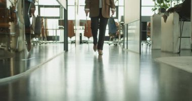 Modern bir ofis koridorunda elinde belgelerle dolaşan bir iş kadınının alt yarısı yakın plan görüntüsü. Gösterişli iç ve cam duvarlar çağdaş bir çalışma ortamını vurguluyor.