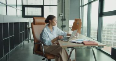 Mavi gömlekli genç bir kadın, elinde bir bardak suyla modern bir ofis masasında çalışıyor. Etrafı çizelgelerle, belgelerle ve elektronik cihazlarla çevrili, büyük pencereli bir ofiste.