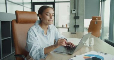 Günümüz ofis ortamında akıllı telefonunu kontrol eden genç profesyonel bir kadın dizüstü bilgisayarıyla çalışıyor. İş haritaları, belgeler ve parlak bir iç mekan verimli bir çalışma ortamını vurguluyor.