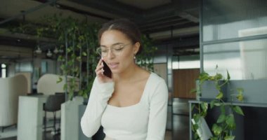 Neşeli, profesyonel bir kadın modern dekorasyon ve gür bitkilerle şık bir ofiste telefonda konuşuyor. Onun kendine güveni ve doğal aydınlatması canlı ve ilham verici bir çalışma alanı ortamı yaratır..