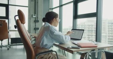 Çağdaş bir ofis alanında çalışan genç profesyonel bir kadın, şehir binalarının manzarasını sunan büyük pencerelerle çevrili mimari planlarını gösteren dizüstü bilgisayarına odaklanıyor.