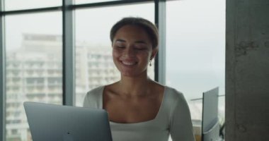 Pürüzsüz ve çağdaş ofis odasında dizüstü bilgisayar kullanan neşeli, genç bir kadın. Büyük camlardan içeri ışık saçıyor ve üretken ve pozitif bir ortam yaratıyor.