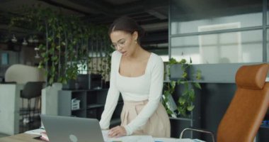 Modern ofis ortamında dizüstü bilgisayarı üzerinde çalışan kararlı genç bir kadın. Öne eğilir, grafikleri ve grafikleri analiz eder, odaklanma, zeka ve hırs gösterir..