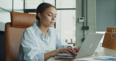 Mavi gömlekli genç profesyonel bir kadın, bilgisayarını modern bir ofis masasında çalışıyor, akıllı telefonunu kullanıyor ve etrafını dokümanlar, çizelgeler ve parlak bir çalışma alanında bir bardak suyla donatıyor.