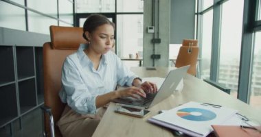Mavi gömlekli neşeli genç bir kadın, modern bir ofis masasında oturuyor. Etrafı çizelgelerle, belgelerle ve geniş pencereli şık bir çalışma alanında akıllı telefonuyla çevrili.