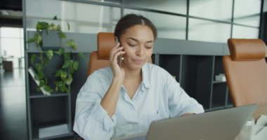 Mavi gömlekli genç profesyonel bir kadın modern bir ofiste masasında oturuyor. Deri koltuklar, bitkiler ve düzenli bir çalışma alanı ile çevrili dizüstü bilgisayarını kullanırken telefonla konuşuyor.