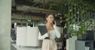  Modern bir ofiste odaklanmış genç bir kadın telefonda konuşuyor ve laptopuyla çalışıyor, yemyeşil bitkiler, rahat mobilyalar ve parlak çağdaş bir iç mekan ile çevrili.