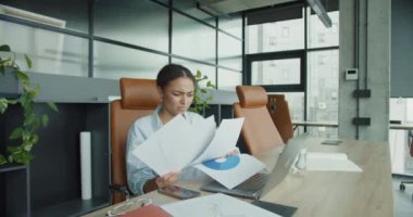 Mavi gömlekli genç profesyonel bir kadın, modern bir ofiste masasında oturmuş, çizelgeler, dizüstü bilgisayarlar ve organize çalışma alanlarıyla çevrili basılı belgeleri inceliyor.