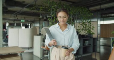 Modern bir ofisteki profesyonel genç bir kadın elinde bir dosya ve kahve fincanı ile gezer, yemyeşil bitkiler, şık iç mekanlar, ve çağdaş mobilyalar, etrafa güven ve odaklanma saçar.