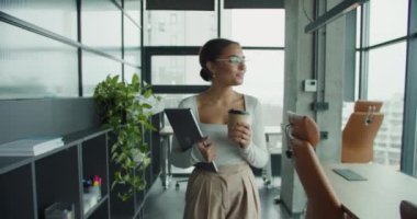 Zarif kıyafetli, elinde bir kahve fincanı ve laptop olan, kendine güvenen genç bir kadın, büyük pencereli, şık mobilyalı ve yeşil bitkilerli, parlak, modern bir ofis alanında yürüyor.