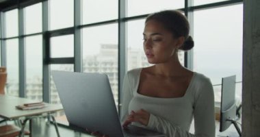 Modern bir ofiste oturan profesyonel bir kadın, dizüstü bilgisayarda yazarken doğal ışık yerden tavana cam pencerelerden geçiyor, şehir manzarasına tepeden bakıyor. 