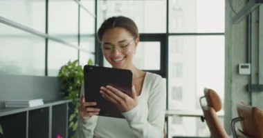 Gülümseyen, gözlüklü, beyaz bluzlu, elinde tablet bir aletle modern bir ofis odasında bitkiler ve parlak mobilyalarla duran profesyonel bir kadın.