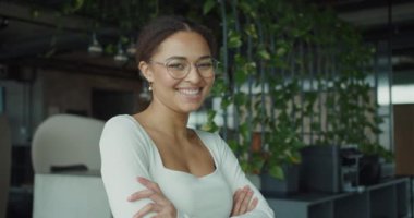 Şık bir ofis ortamında, kollarını kavuşturup gülümseyen neşeli genç bir kadın. Yeşillik ve modern tasarım yaratıcı ve güçlendirici bir çalışma ortamını vurguluyor