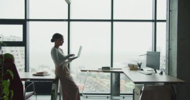 Modern bir ofiste masa başında dizüstü bilgisayarda yazan profesyonel bir genç kadın. Büyük cam pencereler sakin bir çalışma alanını vurgulayarak manzaralı bir kentsel ve kıyı arkaplanı sağlıyor