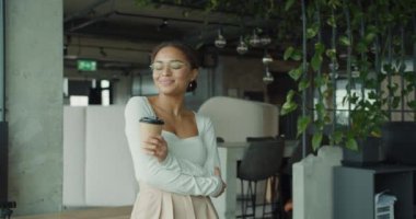 Çağdaş bir ofis ortamında elinde kahve fincanı tutan ve içten gülümseyen neşeli genç bir profesyonel. Canlı, yeşillik dolu çalışma alanı modern ve rahat bir çalışma ortamını vurguluyor.