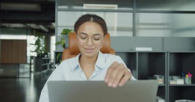 Gözlüklü genç bir kadın modern bir masada şık bir ofiste oturuyor, işini bitirdikten sonra laptopunu kapatıyor, etrafını raflar, bitkiler ve rahatlatıcı bir çalışma ortamı sarmış.