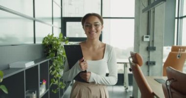 Kendine güvenen, gözlük takan ve elinde bir pano olan genç bir kadın şık deri koltuklar, büyük pencereler ve arka planda yeşillik olan parlak ve modern bir ofis ortamında duruyor.