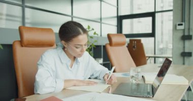 Gözlüklü ve mavi gömlekli genç profesyonel bir kadın modern bir ofis masasında oturuyor, odaklanmış bir şekilde defterine yazıyor, etrafı bir dizüstü bilgisayar, belgeler ve yeşil bitkilerle çevrili, parlak bir çalışma alanında.