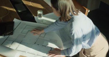 Bir mimarın, dizüstü bilgisayar, gözlük ve bir bardak su gibi gerekli aletlerle doğal ışık altında planları incelediği, üretken bir ortam yaratan profesyonel bir çalışma alanı..