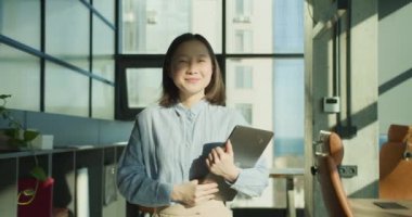 Mavi gömlekli genç bir kadın, elinde dizüstü bilgisayarla modern bir ofiste güvenle duruyor. Geniş pencereler, kentsel ufuk çizgisini ve çalışma alanını vurgulayarak, doğal ışığın boşluğu doldurmasına olanak sağlıyor.