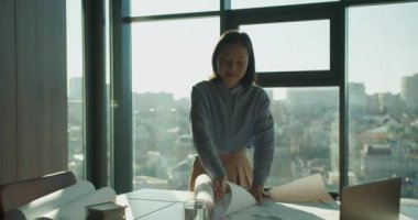 Profesyonel bir mimar, geniş pencereli modern bir ofiste mimari planları dikkatlice inceliyor. Ayar, yaratıcılığı vurgulayan doğal güneş ışığı, şehir görüşleri ve tasarım araçlarına sahiptir.