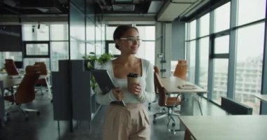 Gözlük takan, elinde bir kahve fincanı ve dizüstü bilgisayarla, geniş pencereli, doğal ışığı olan ve arka planda şehir manzarası olan modern bir ofiste yürüyen kendinden emin genç bir kadın.