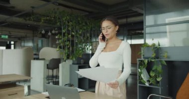 Odaklanmış bir iş kadını modern bir ofiste duruyor, belgeleri incelerken bir telefonla birden fazla iş yapıyor. Çalışma alanı şık, bitkilerle dolu, doğal ışık ve düzenli bir atmosfer..